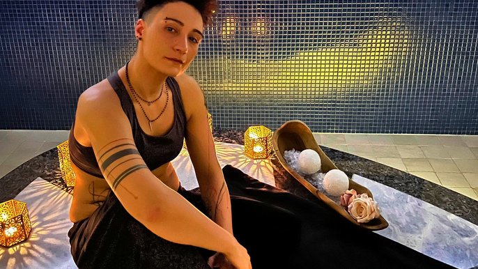 Saunamistrzyni Karolina Grzelak, brunetka o krótkich włosach pozująca na podłodze, na niebieskim tle. Ubrana w czarny krótki top i czarne spodnie. Pozuje obok akcesoriów saunowych i świec. Na prawym ramieniu ma tatuaż. 