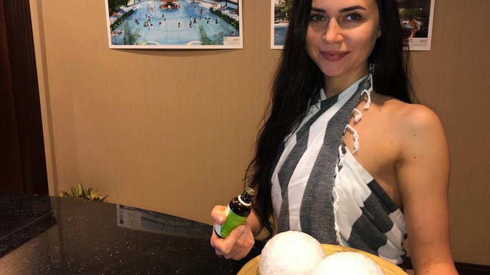 Saunamistrzyni Sylwia Skarbek. Uśmiechnięta młoda brunetka z długimi włosami trzyma w jednej dłoni fiolkę z olejkiem eterycznym, a w drugiej tacę z dwiema lodowymi kulami. Ma na sobie hammam w biało-szare pasy. 