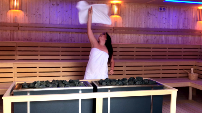 Wnętrze dużej sauny zewnętrznej. Na pierwszym planie grafitowy piec saunowy z drewnianą ramą, a za nim saunamistrzyni Sylwia Drobiszewska owinięta białym ręcznikiem kręci nad głową drugim ręcznikiem niczym pizzą. 