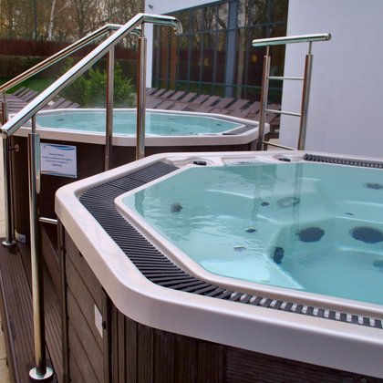 Dwie wanny jacuzzi obudowane ciemnym drewnem, pomiędzy nimi podest z aluminiową poręczą. W tle plaża terenu zewnętrznego Strefy Saun.