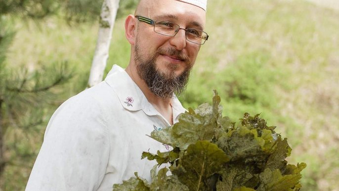 Saunamistrz Marek Olszewski ma na sobie białą koszulę i nakrycie głowy, na nosie okulary, a w rękach trzyma witki brzozowe. 