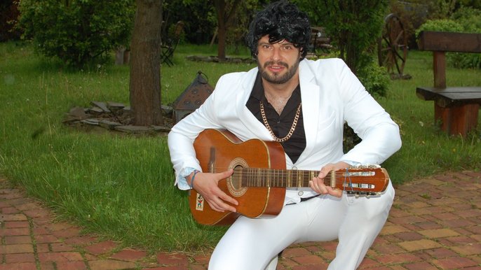 Saunamistrz Łukasz Gapiński, mężczyzna z ciemnym zarostem, w białym garniturze i czarnej koszuli pozuje na tle drzew. Pozuje kucając. Trzyma gitarę. Na głowie ma perukę z czarnymi kręconymi włosami. Na szyi ma złoty łańcuszek. 