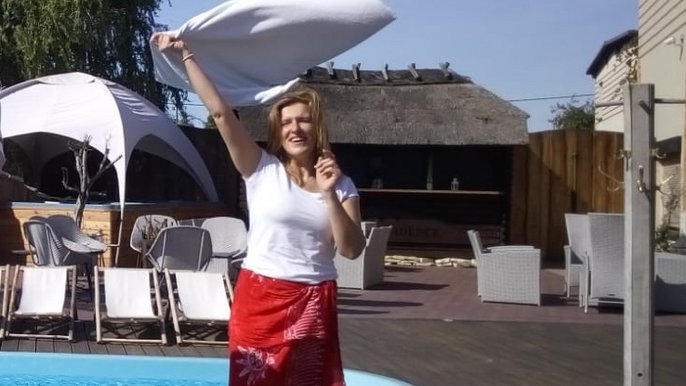 Szeroko uśmiechnięta blondynka w białej koszulce i czerwonym hamamie kręci ręcznikiem nad głową trzymając go w uniesionej prawej dłoni. W tle basen i białe leżaki. 