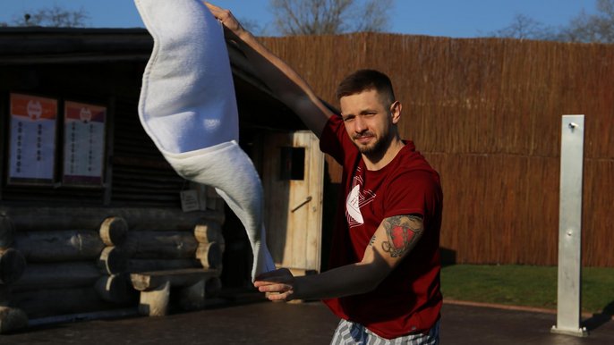 Młody mężczyzna z lekkim zarostem ma na sobie bordową koszulkę i hammam w paski. W wyciągniętych przed siebie rękach trzyma rozłożony szeroko ręcznik. 