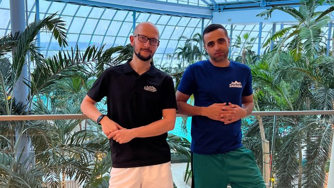Saunamistrzowie Przemek Sawicki (w okularach od lewej) i Hatem Mhadhbi (od prawej). Mężczyźni pozują na tle palm w parku wodnym SUNTAGO. Przemek ma na sobie czarny t-shirt i białe spodenki. Hatem ma granatowy t-shirt i zielone spodenki. 