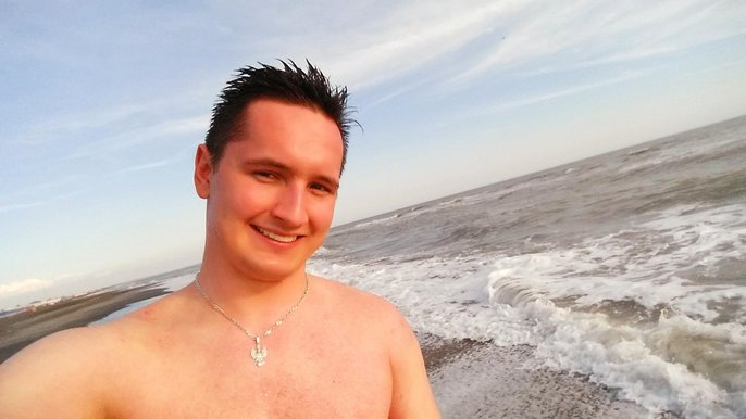 Uśmiechnięty mężczyzna o krótkich, ciemnych włosach pozuje na tle morza. Ujęcie portretowe ukazuje mężczyznę od wysokości barków w górę. Morze w tle lekko wzburzone. 