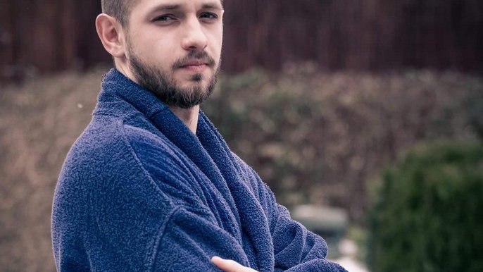 Saunamistrz Michał Blejder. Młody brunet z zarostem ubrany jest w granatowy szlafrok i pozuje na tle terenu zewnętrznego Strefy Saun. Ręce ma splecione na piersi, stoi bokiem do obiektywu. 