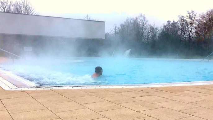 Zewnętrzny basen wypływowy w zimowy dzień - jeden z Falowiczów korzysta z uroków wodno-powietrznych leżaków, w tle gęsta para unosząca się nad wodą i hala z basenem sportowym. 