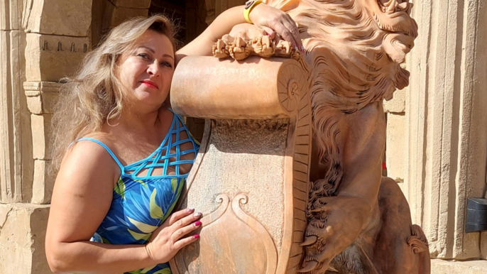 Saunamistrzyni Mirela Kazuś-Barańska. Blondynka we wzorzystej, niebieskiej sukience na ramiączka. Pozuje oparta o figurę lwa wykonaną z kamienia w kolorze jasnego brązu. W tle jest widoczny zabytkowy budynek również w tym kolorze. 