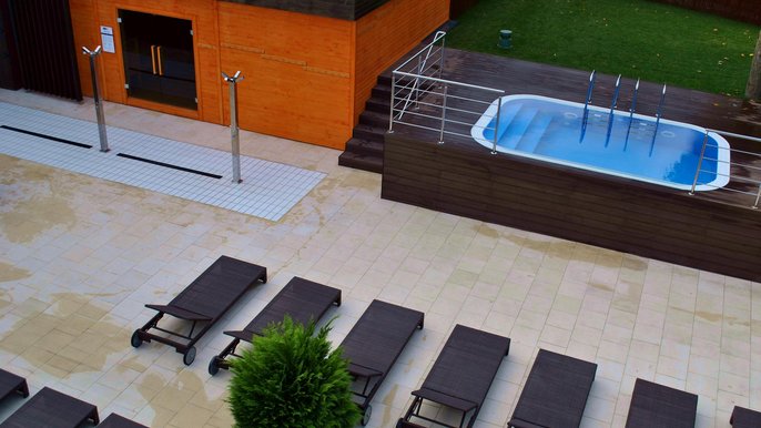 Panorama terenu zewnętrznego Strefy Saun. Od lewej duża sauna zewnętrzna, dalej basen wody zimnej i kawałek zielonego trawnika, a poniżej plaża z rzędami ciemnych ręczników. - Panorama terenu zewnętrznego Strefy Saun. Od lewej duża sauna zewnętrzna, dalej basen wody zimnej i kawałek zielonego trawnika, a poniżej plaża z rzędami ciemnych ręczników.