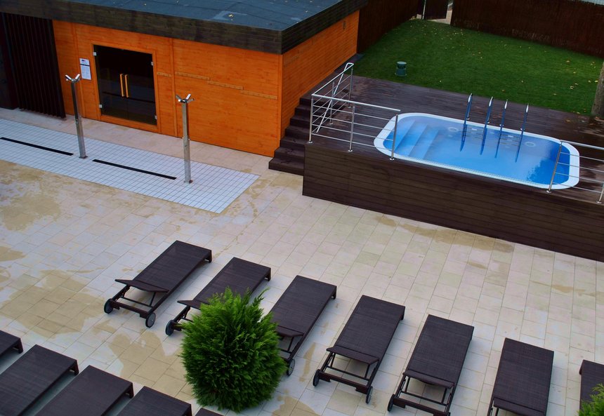 Panorama terenu zewnętrznego Strefy Saun. Od lewej duża sauna zewnętrzna, dalej basen wody zimnej i kawałek zielonego trawnika, a poniżej plaża z rzędami ciemnych ręczników.