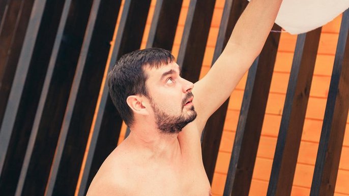 Saunamistrz Damian Rurarz. Brunet z zadbanym zarostem na uniesionej nad głową ręce utrzymuje płasko rozłożony ręcznik. W drugiej dłoni trzyma drugi biały ręcznik. 
