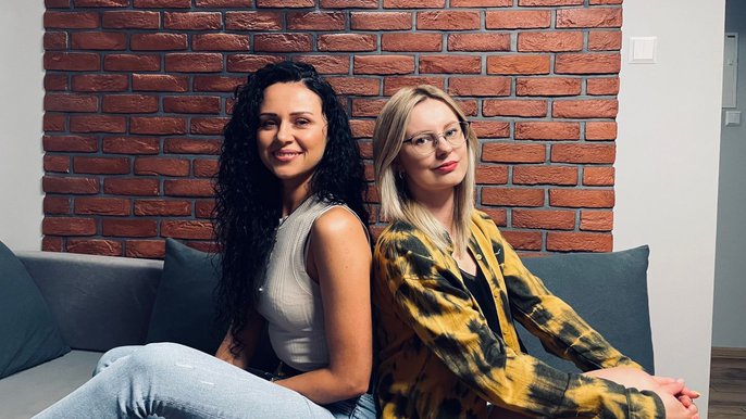 Z lewej strony saunamistrzyni Sylwia Skarbek w szarej bluzce bez rękawów i niebieskich spodniach. Z prawej strony saunamistrzyni Marlena Wolder w żółto zielonej sukience z długim rękawem. Kobiety siedzą i opierają się o siebie plecami. 