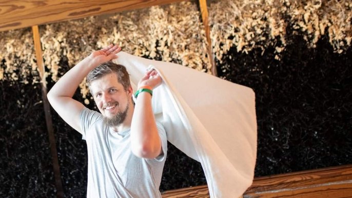 Maciej Kucharski, uśmiechnięty ciemny blondyn z brodą. Ubrany w szary t-shirt. W tle widać witki z tarniny. Mężczyzna na zdjęciu macha ręcznikiem. 