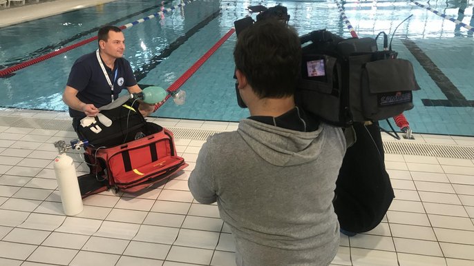 Przy krawędzi basenu sportowego przełożony ratowników WOPR Aquaparku FALA prezentuje wyposażenie czerwonej torby ratowniczej, koło niego dziennikarz, a wszystko dokumentuje stojący tyłem do obiektywu operator z kamerą telewizyjną. 